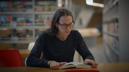 A focused student is deeply engaged in reading a book in the library while wearing glasses