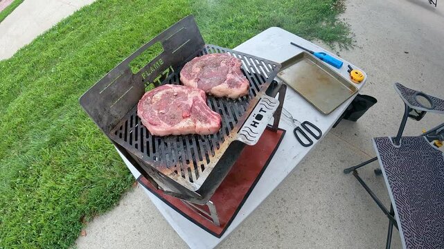 Top view of two ribeye steaks recently placed on a hot charcoal, portable and collapsible grill; concepts of tailgates, picnics and camping