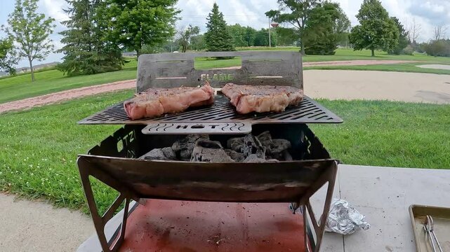 Pan down on portable charcoal grill cooking two thick ribeye steaks; concepts tailgate, picnic, and camping