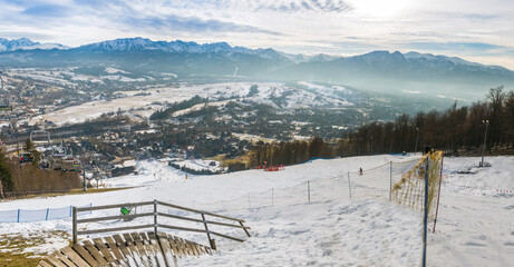 Obraz premium Snowy slopes in Zakopane, Poland, teem with skiers and snowboarders, descending towards a valley lined with scattered buildings