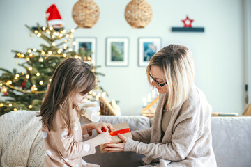 A woman and her daughter are seated on a couch, a girl is opening a holiday gift, as a Christmas tree glows in the background