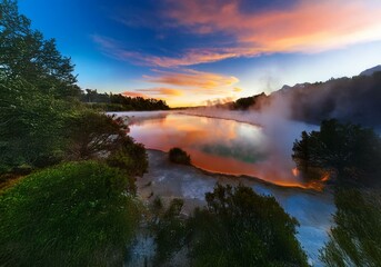 A vibrant New Zealand landscape with a fiery sunrise reflected in a steamy lake.