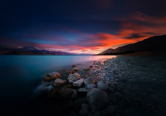 Fototapeta premium A serene New Zealand landscape featuring a still lake, a rocky shoreline, and a vibrant sunset sky.