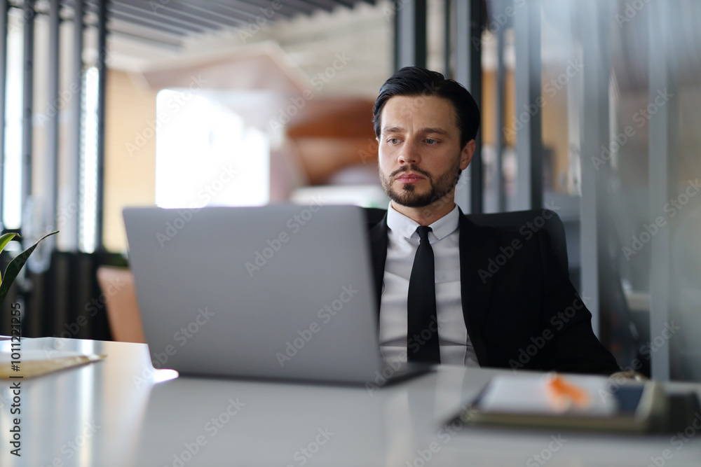 Poster a man in a suit is sitting at a desk with a laptop open in front of him. he is focused on his work, 