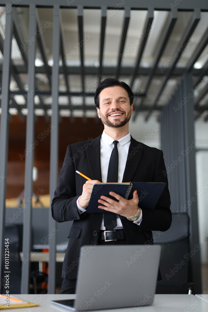 Poster a man in a suit is smiling and holding a notebook and a pen. he is standing in front of a laptop and