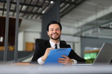 A man in a suit is smiling and holding a blue clip board. He is sitting at a desk with a laptop in front of him