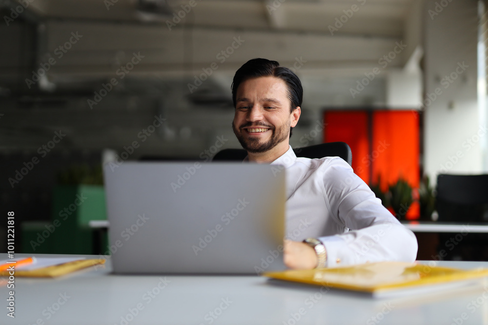 Poster a man is sitting at a desk with a laptop open in front of him. he is smiling and he is enjoying his 