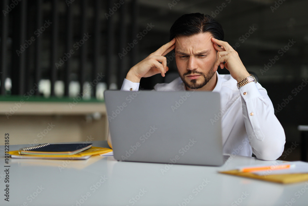 Poster a man is sitting at a desk with a laptop open in front of him. he is looking at the screen with a pu