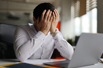 A man is sitting at a desk with a laptop and a stack of papers. He is crying and he is in distress