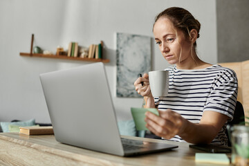 A woman with vitiligo sips coffee, focused on her laptop screen during a cozy morning.