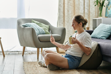 A young woman relaxes on the floor, sipping tea while engaging with her smartphone.