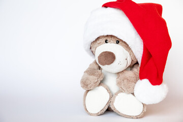 Cute teddy bear toy with smile in huge Santa hat sits on white background where there is copy space