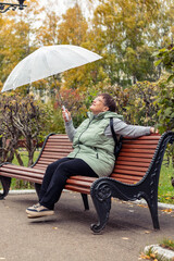 An elderly woman with an umbrella is sitting on a bench.