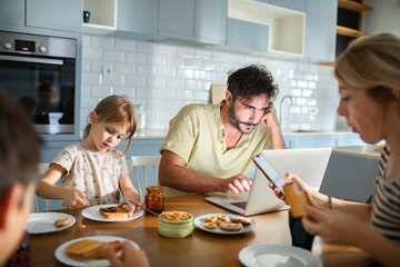 Father working on laptop while family has breakfast in kitchen