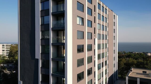 A drone flyby showcasing a modern high-rise building with sleek balconies and large windows. The ocean and surrounding greenery in the background enhance the urban-coastal appeal