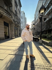 Millennial man stands confidently on a sunlit city street, wearing a cozy jacket and stylish hat....