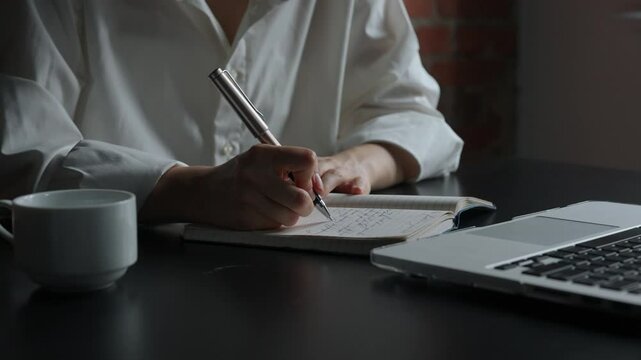 Business woman sitting at office desk with laptop writing notes in dairy notebook with pen, concentrated person writing down ideas and thoughts, close up shot.