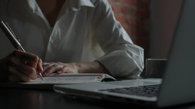 Business woman sitting at office desk with laptop writing notes in dairy notebook with pen, concentrated person writing down ideas and thoughts, close up shot.