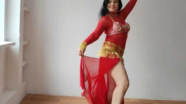A dancer in a red suit is dancing an oriental dance on a white background. A professional dancer dances oriental belly dance. Sexy woman in red dress dances a seductive dance in a white studio