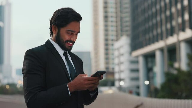 Young businessman scrolling through mobile telephone and reading pleasant news at empty city street. Relaxed male checking mailbox and noticing long-waited invitation for meeting at evening.
