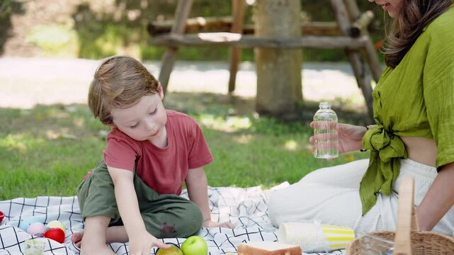 Caucasian mother, looking at son sitting in front, with loving eyes, boy puts bread on plate, before reaching out grabbing another piece dessert from plate eat, have picnic in garden.