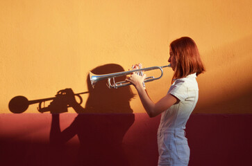 A young girl is playing a trumpet in front of orangewall. 