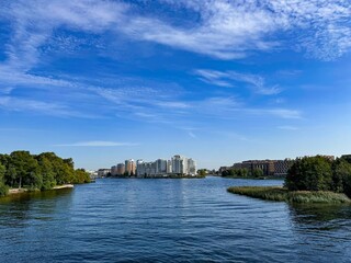 A peaceful riverside scene featuring calm water, lush greenery on the banks, and modern residential buildings in the distance. The clear blue sky with light clouds adds to the serenity of this urban