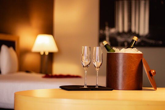A romantic hotel room setting with a chilled bottle of champagne and two glasses on a table, adorned with rose petals on the bed in the background. Valentine's Day, anniversaries, or honeymoon