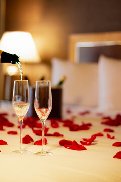A romantic hotel room setting with a chilled bottle of champagne and two glasses on a table, adorned with rose petals on the bed in the background. Valentine's Day, anniversaries, or honeymoon