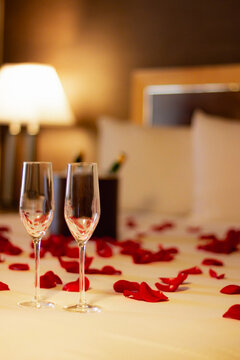 A romantic hotel room setting with a chilled bottle of champagne and two glasses on a table, adorned with rose petals on the bed in the background. Valentine's Day, anniversaries, or honeymoon