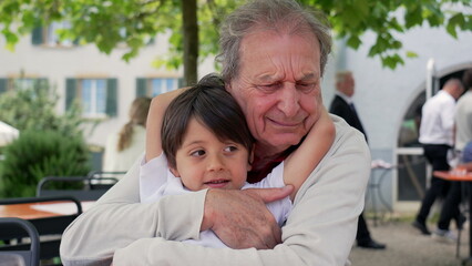 Grandfather holding his grandson at an outdoor cafe, both smiling and enjoying each other's company, surrounded by greenery, capturing a warm and loving family moment