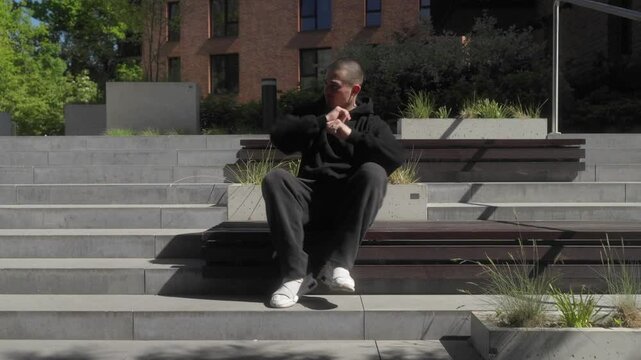 A man in a hoodie sits on stairs in a modern city setting, energetically dancing and gesturing with his hands and feet. The relaxed yet dynamic scene reflects youth, confidence, and urban style.