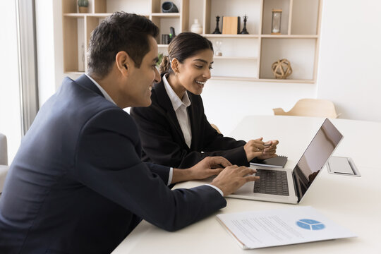 Experienced Indian male employee manager executive mentoring coaching female intern trainee on workplace teach to use corporate software. Diverse coworkers cooperate prepare document report on laptop