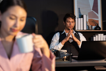 Focusing on sitting man coworker working at back desk view while waiting email business project report on laptop at late night time with blurry woman background drinking coffee at office. Postulate.