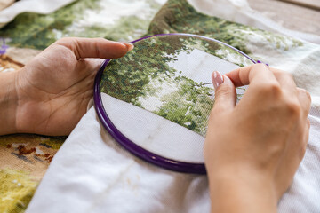 A girl cross-stitching. Close-up of hands carefully maneuvering a needle through the fabric. A purple embroidery hoop holds the fabric. Close-up of hands with a needle threading the fabric.