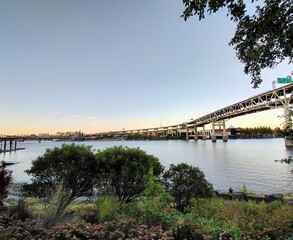 The image captures the Portland road bridge spanning across the calm waters of a river during a serene summer evening in 2023. The sky is clear with soft, warm hues as the sun sets.