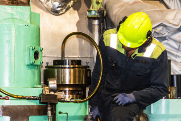 A close-up of the upper body and head of a marine engineer officer, wearing a helmet, focused on performing duties in the engine room during repair work of the ship’s main engine.