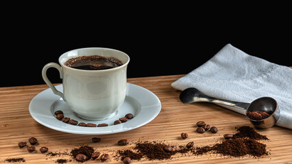 A white cup of coffee, spreaded coffee beans, milled coffee, silver spoon, wooden desk, white wiper and Black background