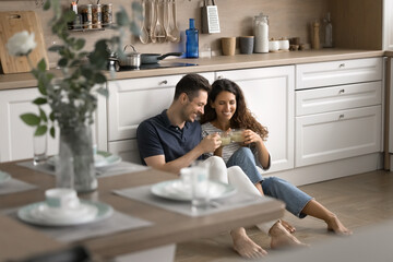 Cheerful positive young romantic couple sitting on heating floor in modern kitchen, toasting mugs...