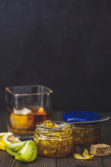 Jars of green tomato jam with a spoon resting on top and a glass of whiskey in the background