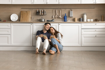 Happy couple of homeowners sitting on floor close together in new spacious kitchen interior,...