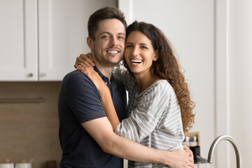 Cheerful young couple in love hugging with faces touch in kitchen, looking at camera with toothy smiles, enjoying domestic romantic leisure time, posing for portrait in home apartment