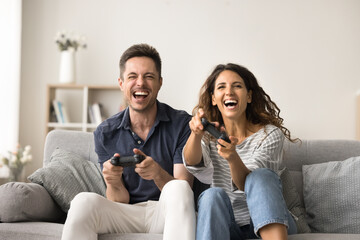 Joyful happy couple excited with video game, virtual entertainment, holding gamepads, pushing buttons of game controllers, shouting, laughing, pushing buttons, sitting on home sofa, front view © fizkes