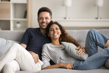 Obraz premium Happy pretty young wife and handsome husband enjoying romantic relationship, leisure on home couch together, hugging with love, affection, looking at camera with toothy smiles