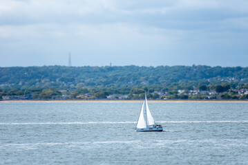 A tourist yacht sails under full sails across the calm Atlantic waters near the coast of the USA, close to New York. The water shimmers silver, reflecting sunlight piercing through thick clouds.