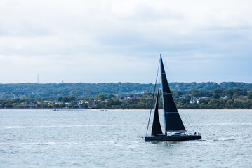 A tourist yacht sails under full sails across the calm Atlantic waters near the coast of the USA, close to New York. The water shimmers silver, reflecting sunlight piercing through thick clouds.