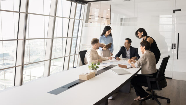 Multiethnic team of happy colleagues and Indian leader discussing business success at meeting table. Diverse employees talking to boss, smiling, laughing, enjoying successful teamwork. Wide shot