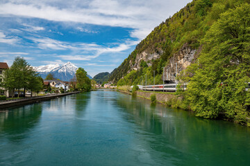 Picturesque view of a turquoise river in Interlaken, with a passing train, lush greenery, charming houses, and snow-capped Alps in the background. Captures the essence of Swiss beauty