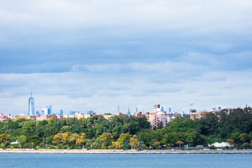 Fototapeta premium The shoreline near the Verrazzano-Narrows Bridge showcases the peaceful Bay Ridge neighborhood, where houses nestle among lush greenery, creating a serene contrast to the bustling city beyond.