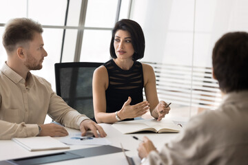 Beautiful young entrepreneur woman talking to male colleagues at office large table, explaining project strategy, speaking to coworkers. Board of directors, business partners meeting for negotiation